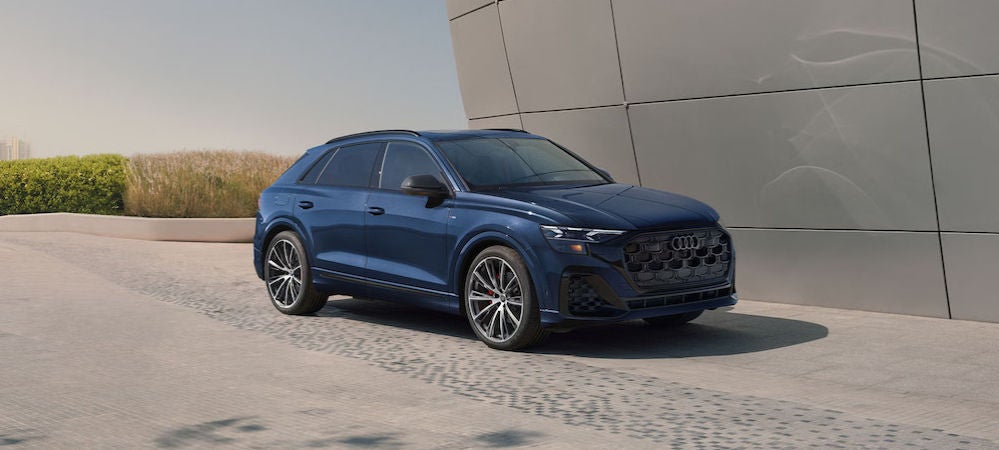 A dark blue 2026 Audi Q8 SUV parked on a driveway in front of a gray modernist building