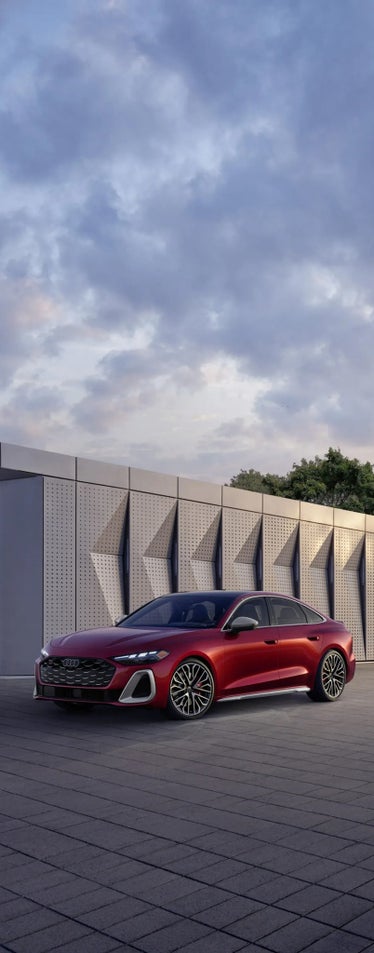 Three-quarter front view of the Audi S5 accelerating past a modern building.