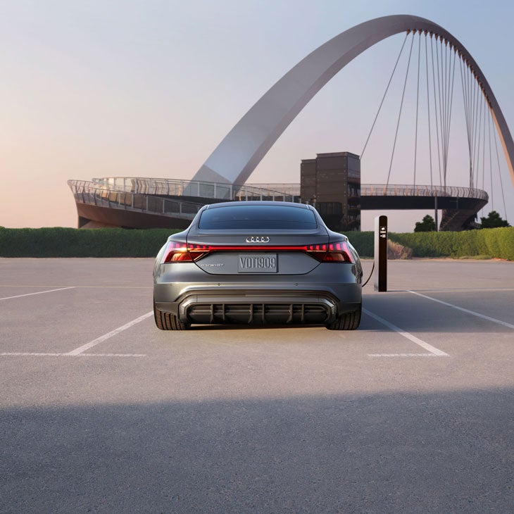 Rear view of a blue Audi e-tron GT charging in front of a futuristic suspension bridge.