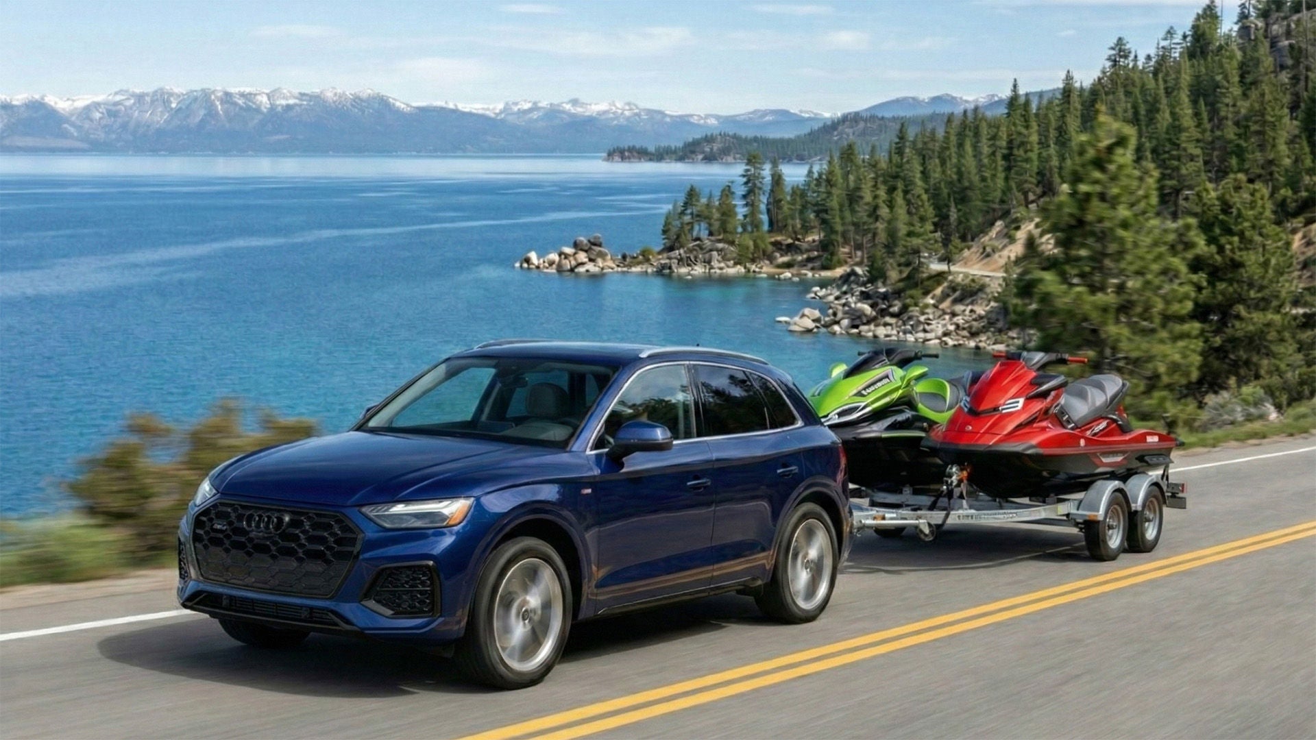 Audi Q5 pulling a trailer of jet skis next to a snow capped mountain lake
