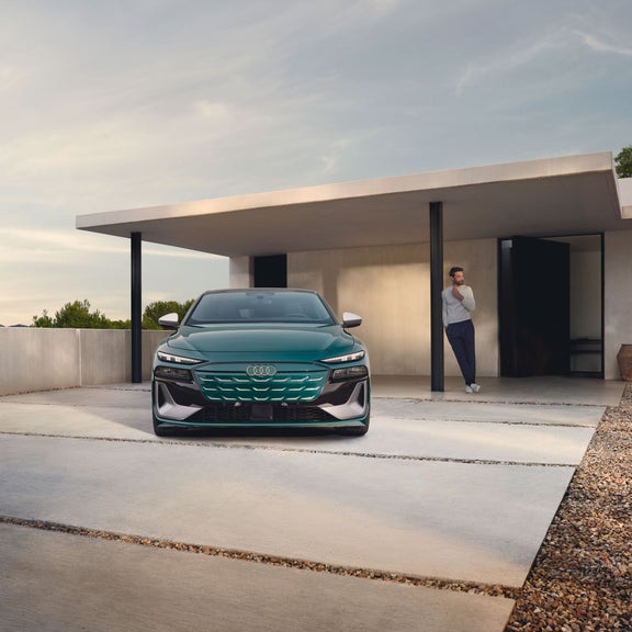 Front view of the Audi S6 e-tron parked in front of a modern home with a person leaning against a pillar nearby.