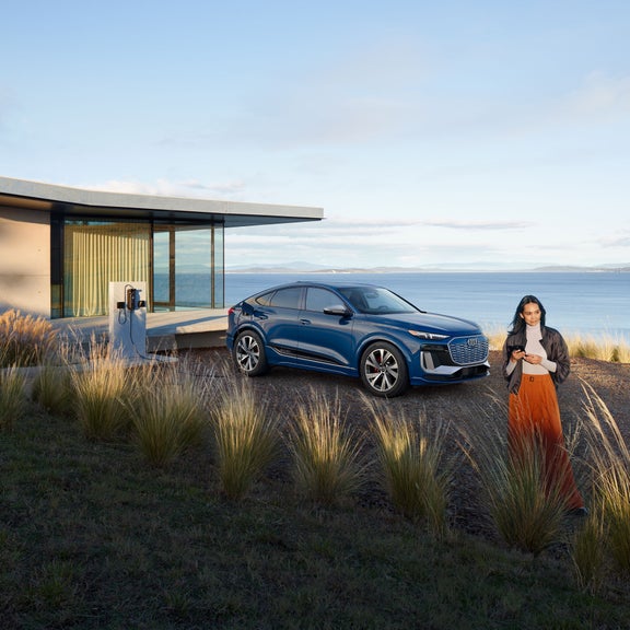 Three-quarter side view of the Audi SQ6 Sportback e-tron parked and charging by a waterfront home with a person standing nearby.