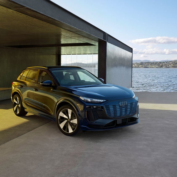 Three-quarter front view of a blue Audi Q6 e-tron parked by a modern building on a waterfront.