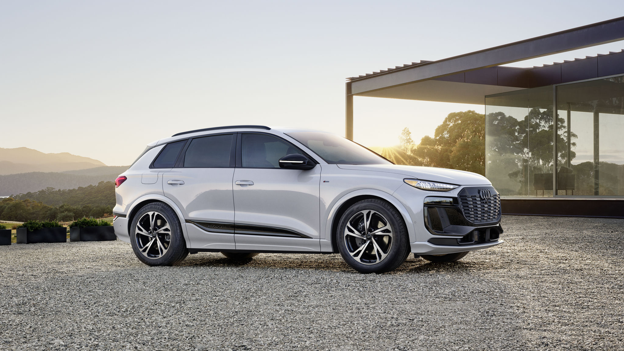 2027 silver Q6 e-tron on gravel with a sunset and mountains in the background.  