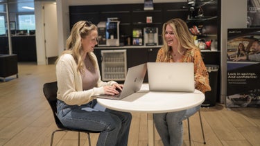 Two women sitting at a small, round table in a waiting room, with snacks and coffee in the background.