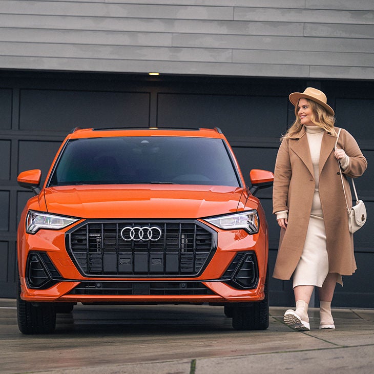 A woman standing next to a parked orange Audi SUV.