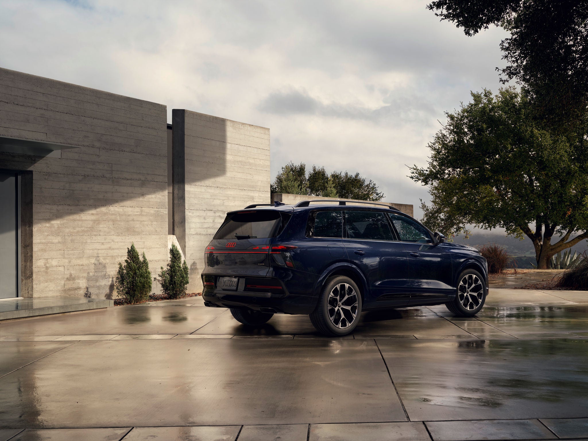Dark blue 2027 Audi Q9 shown from the rear three-quarter angle outside a modern home.