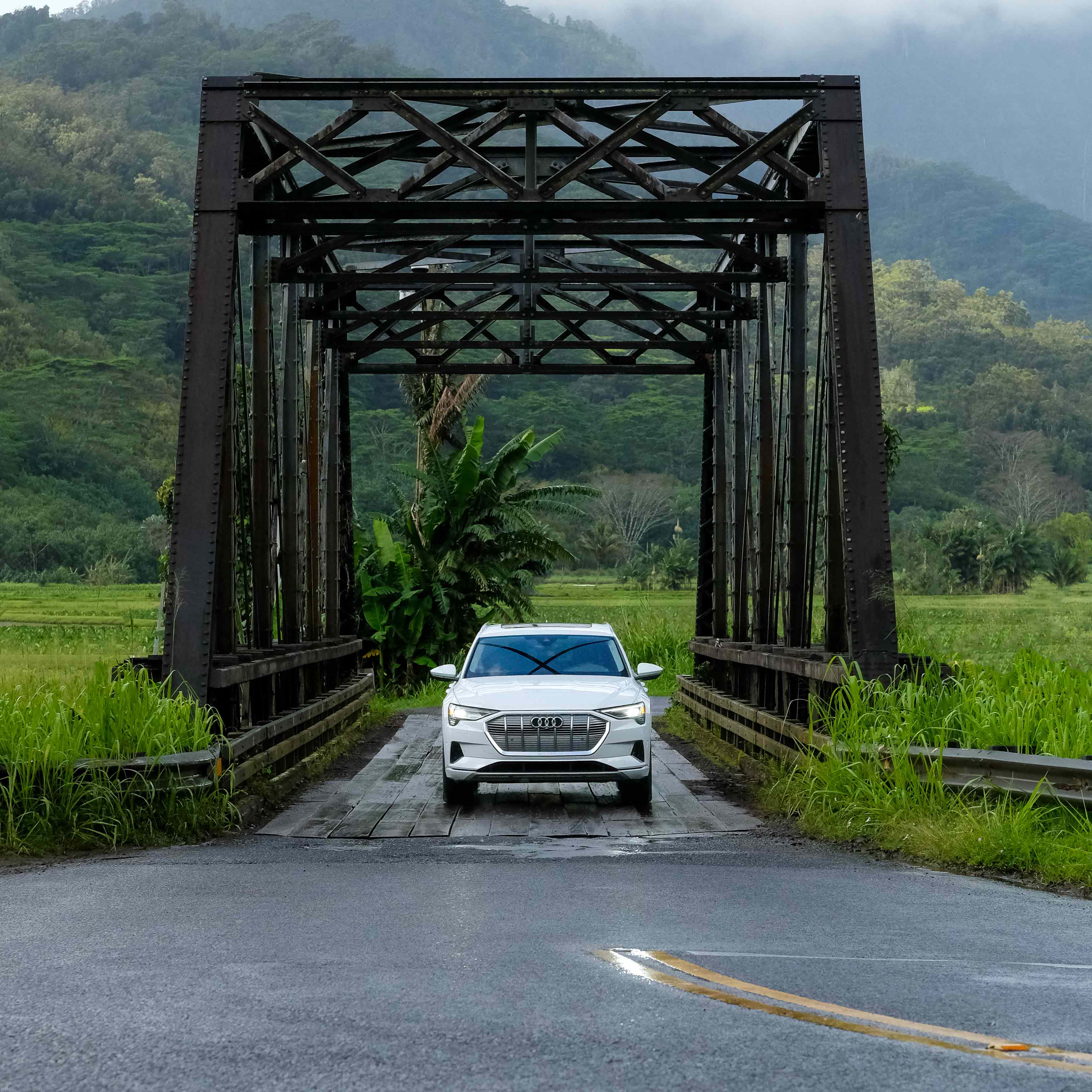 Front view of a white Audi e-tron parked on a bridge in a tropical mountain landscape. 