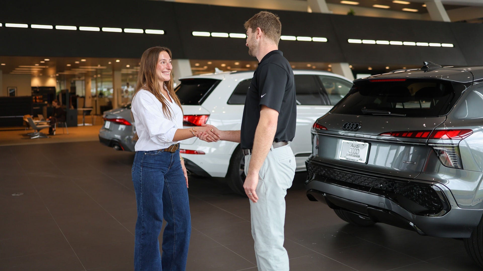 A woman and a man shaking hands inside the Audi Birmingham showroom with a row of cars shown in the background.