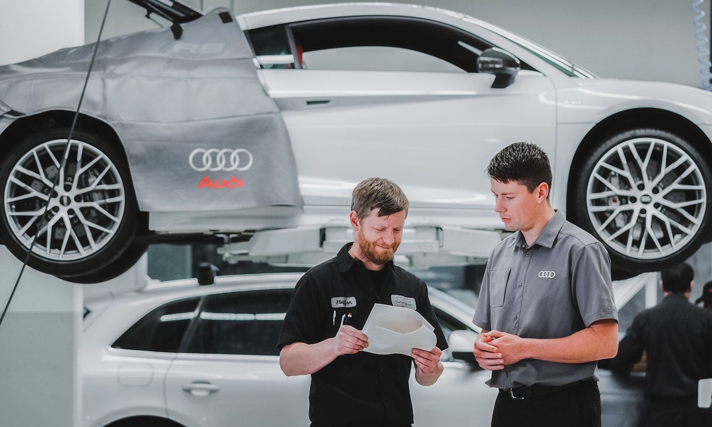 Deux techniciens Audi discutent avec un véhicule Audi suspendu derrière eux.