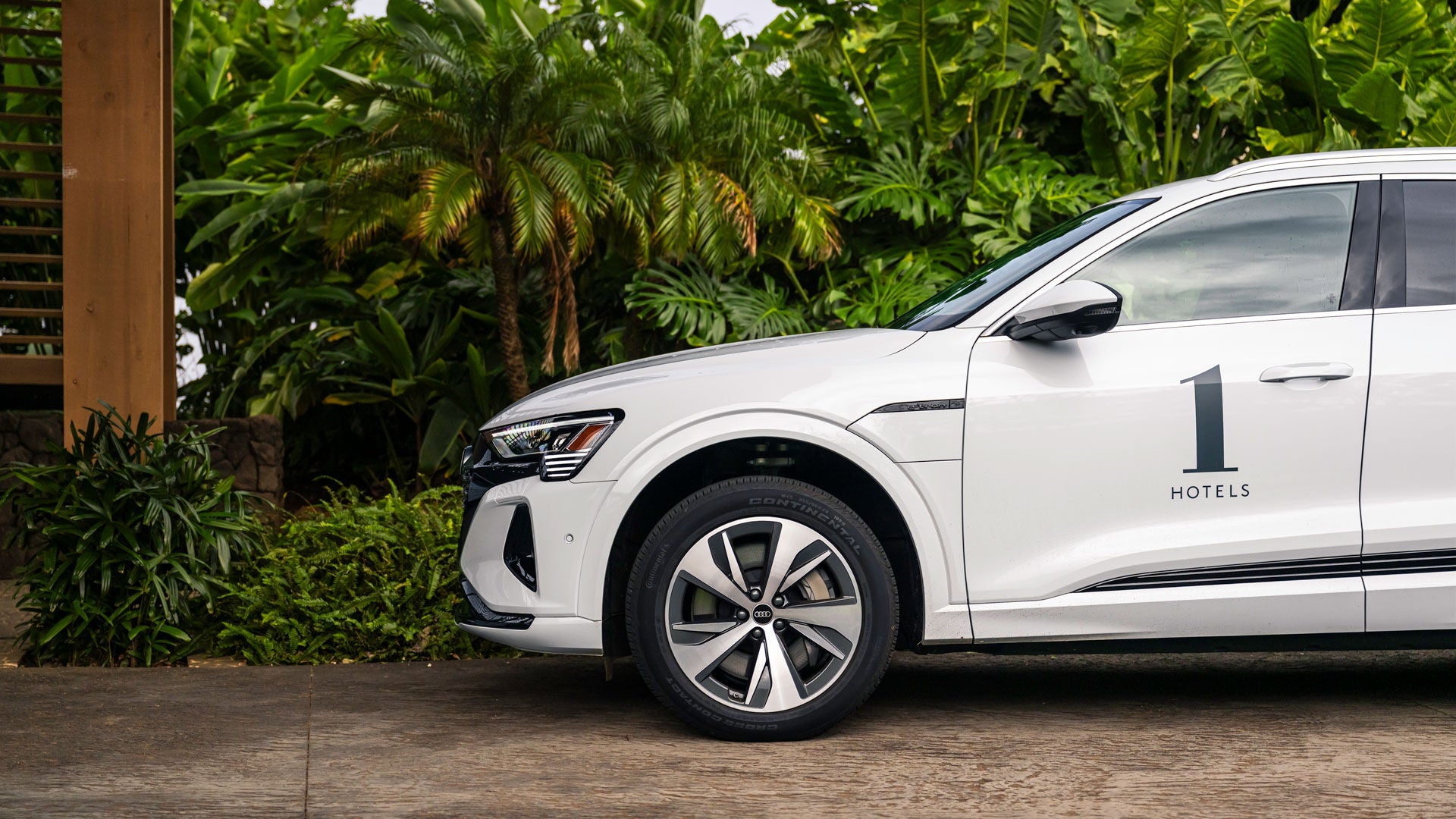 Side profile of a white Audi e-tron with a 1 Hotels logo on it parked in front of an upscale hotel.