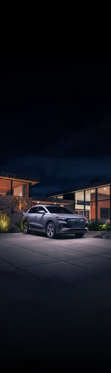 A silver Audi Q8 e-tron charging in front of a modern home at night.