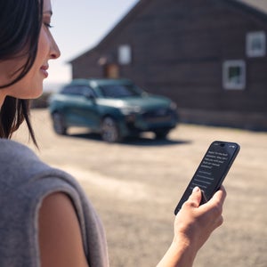 Over-the-shoulder view of a person using the myAudi app with an Audi Q3 parked in the background.