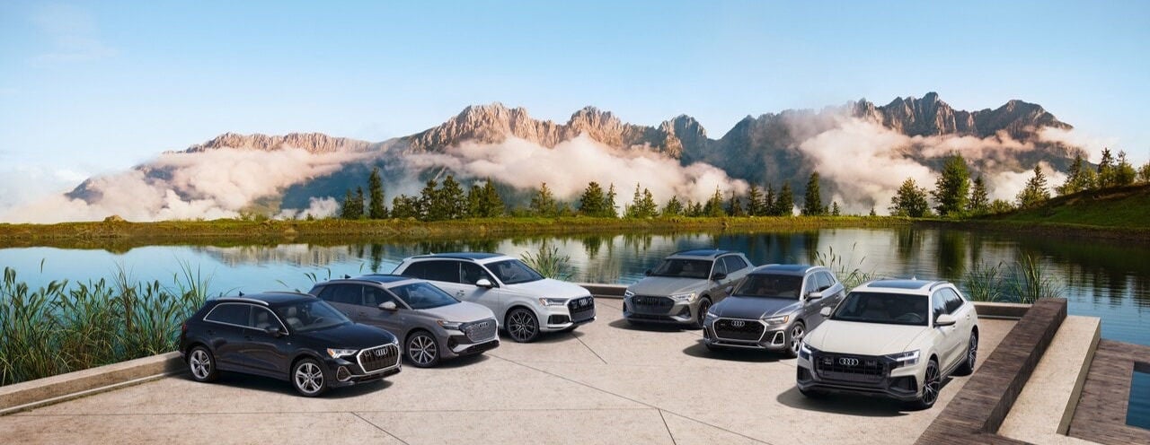 A lineup of Audi vehicles parked beside a scenic mountain lake with snow-capped mountains and clouds in the background.