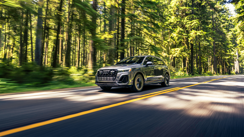 Dark gray Audi Q7 2026 driving on a paved road with green trees in the background during daytime. 
