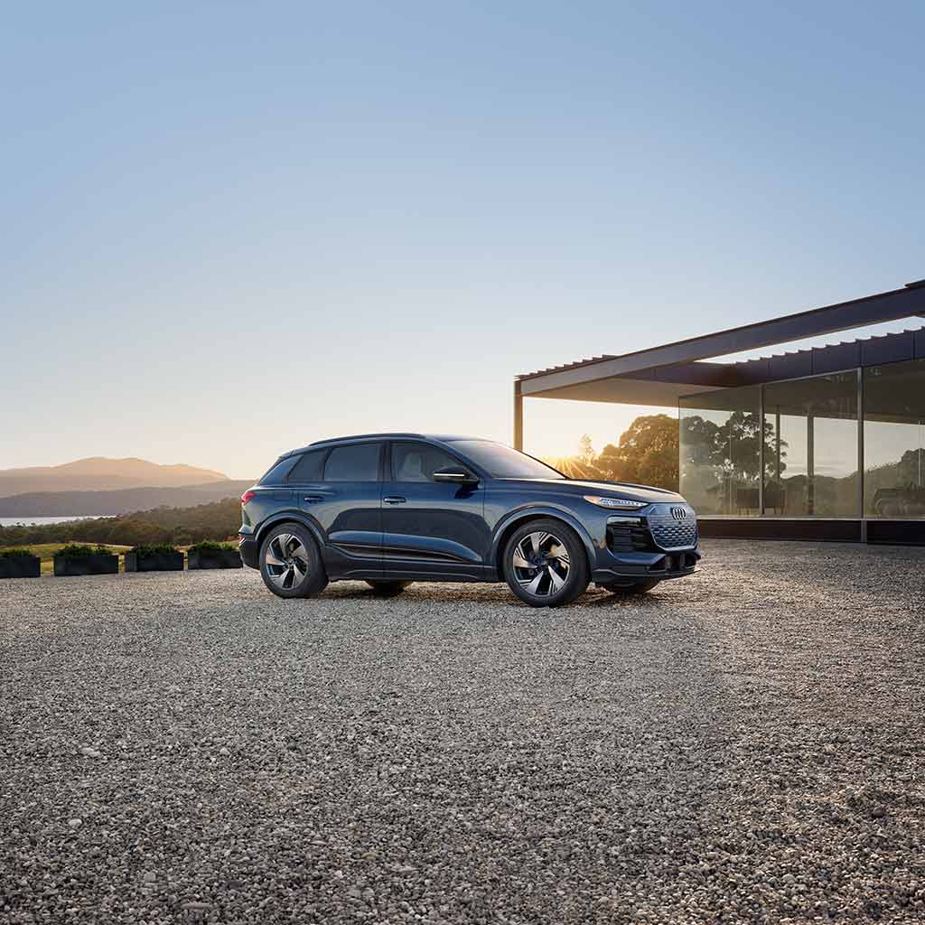 Side profile view of an Audi Q6 e-tron parked outside a modern-looking house.