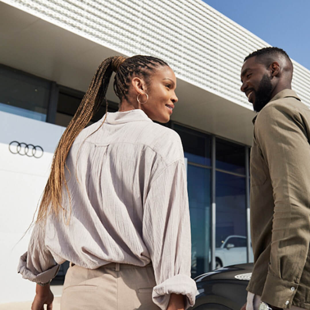 A smiling couple stands outside an Audi dealership in daylight, with the Audi logo and showroom visible behind them.