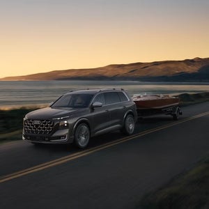 Grey Audi SQ9 towing a boat along a scenic lakeside road at sunset.