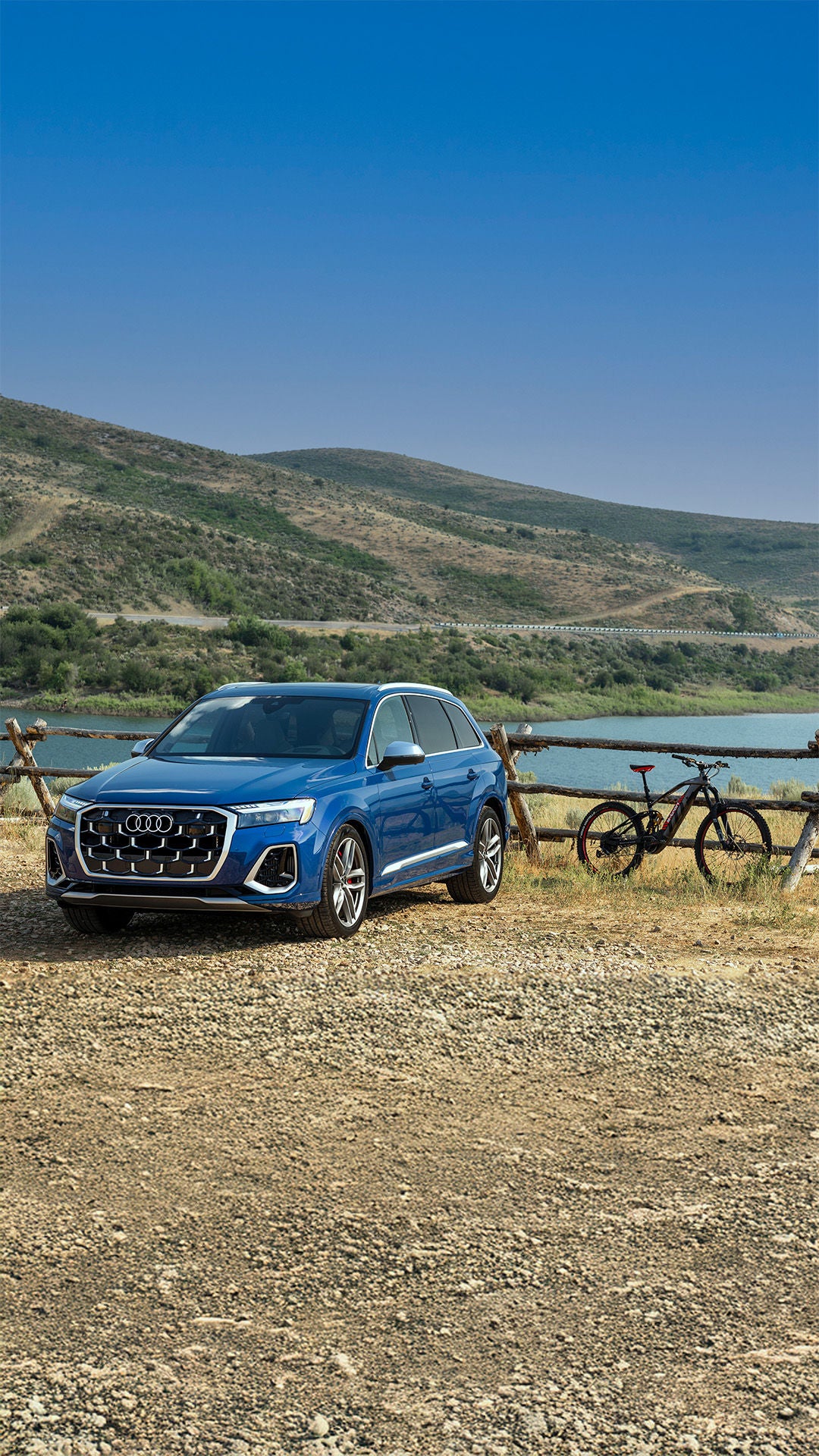 2025 Audi Q7 trim in blue parked above a lake with bike proped on fence and mountains in background.