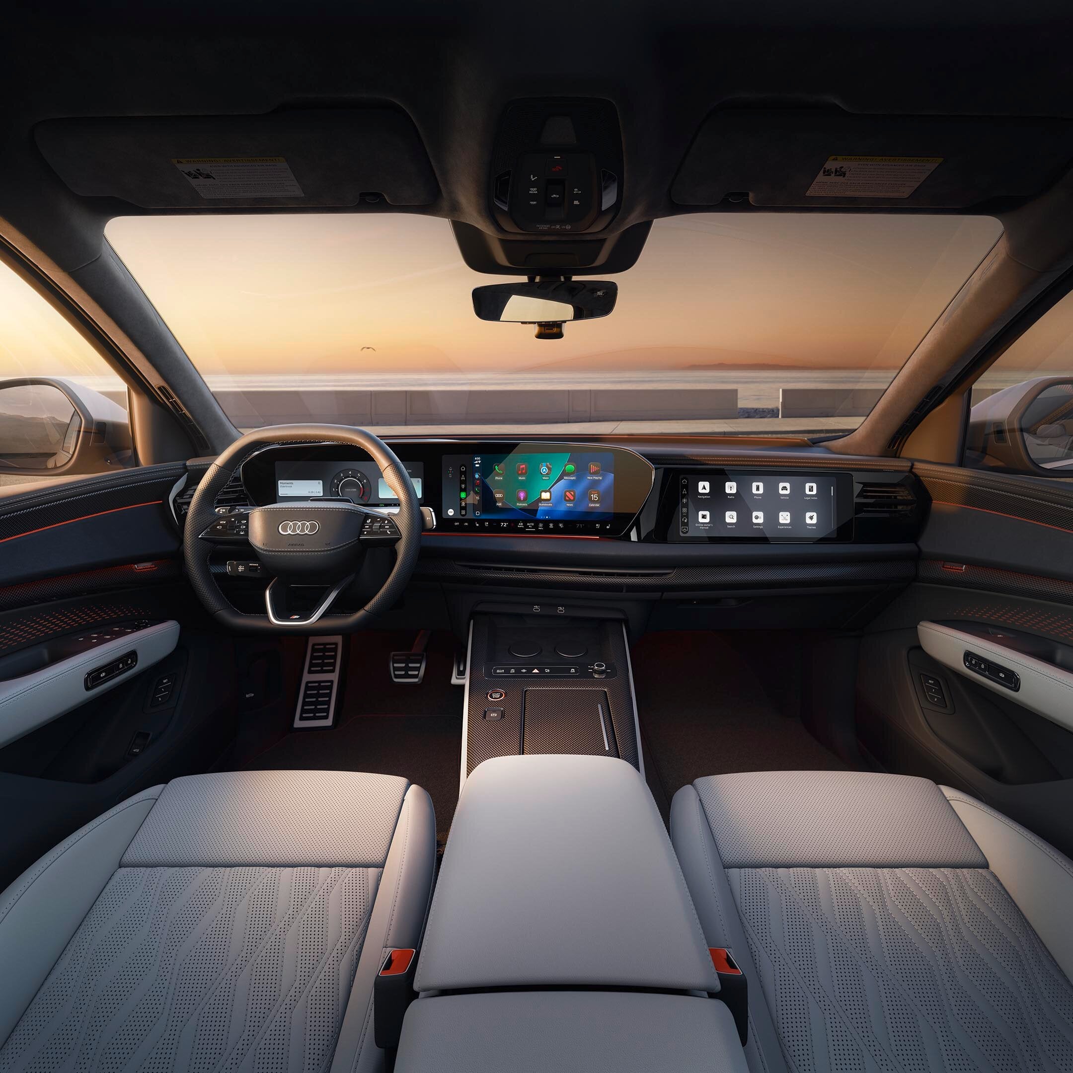 Front interior view of an Audi SQ9 with a two-spoke steering wheel, dual digital displays, and gray leather seats, facing an oceanfront sunset through the windshield.