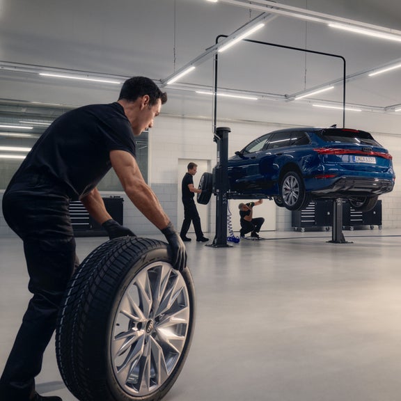 An Audi technician rolling a wheel to a suspended Audi vehicle that is being worked on.