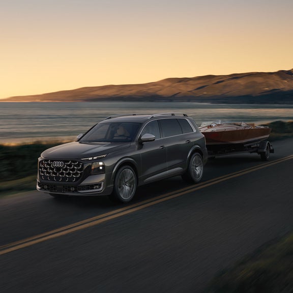 The Audi SQ9 SUV, viewed from the front at an angle on a coastal road, with a motorboat in tow.
