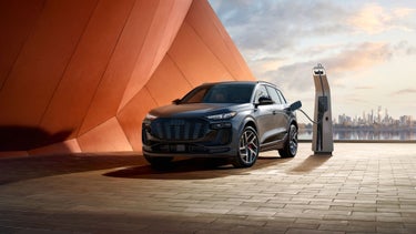 Three-quarter front view of the Audi SQ6 e-tron parked and charging with a cityscape in the background.
