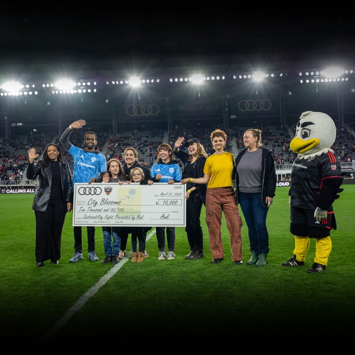 A group of people and a mascot standing on the Audi field, holding a large cheque presented by Audi and D.C. United.