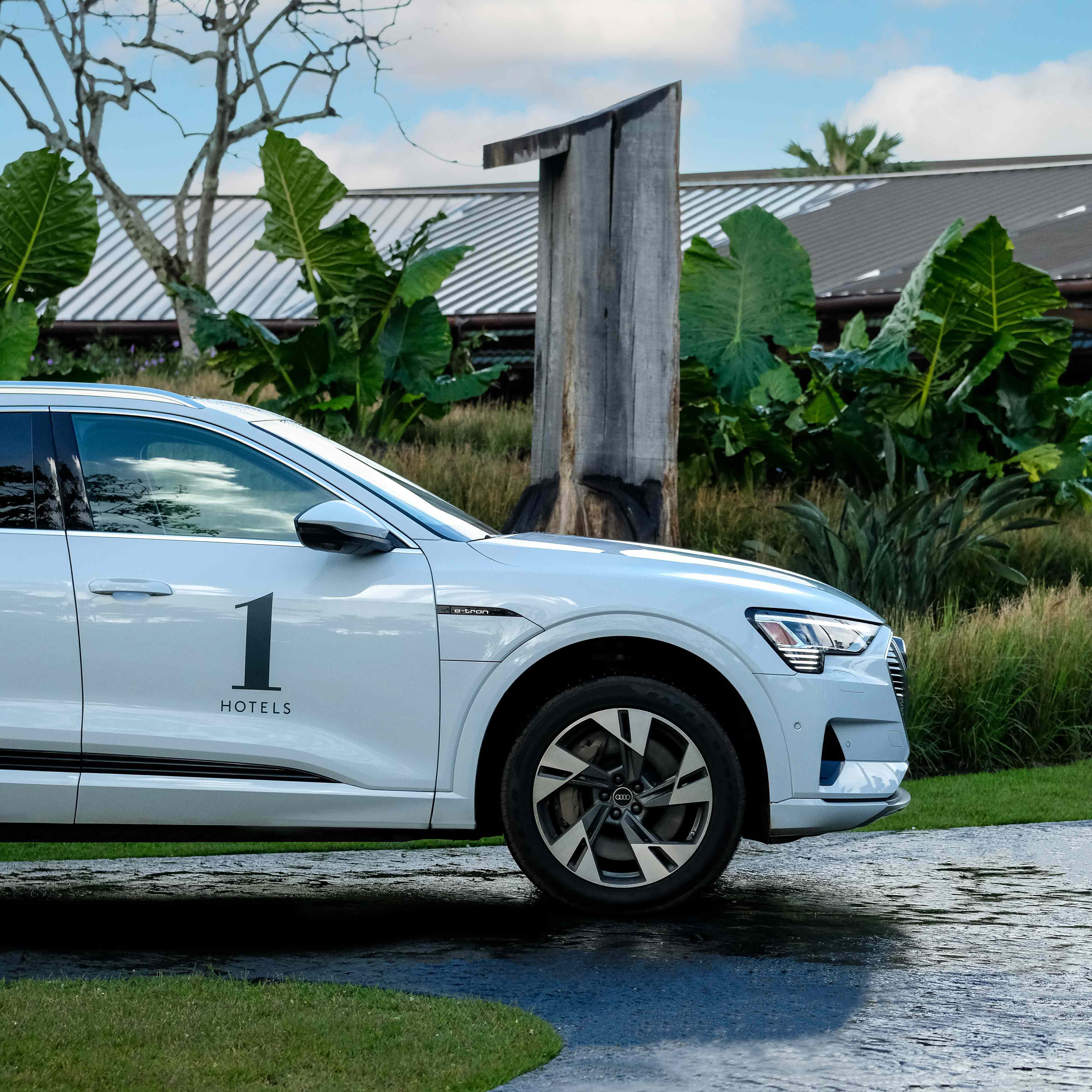 Side profile of a white Audi e-tron with a 1 Hotels logo on it parked in front of a tropical garden.