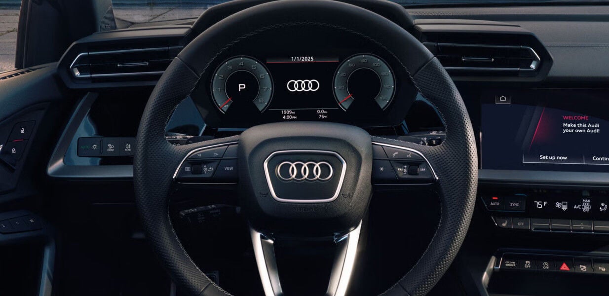 The wheel and driver's side dashboard of the 2026 Audi A3 featuring black leather interior and gray stitching