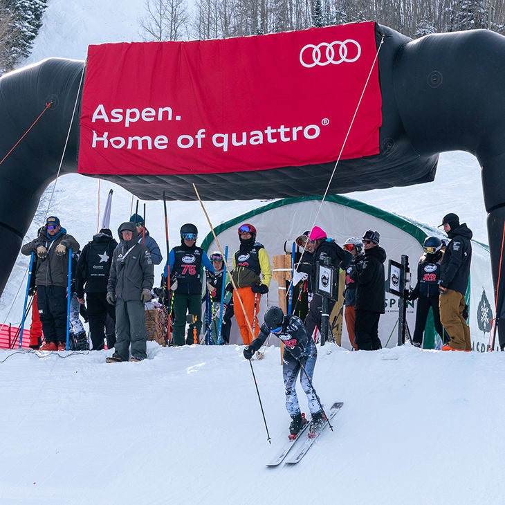 A group of skiers watching a skier start their descent on a snowy hill with an Aspen Home of Quattro banner in the background.