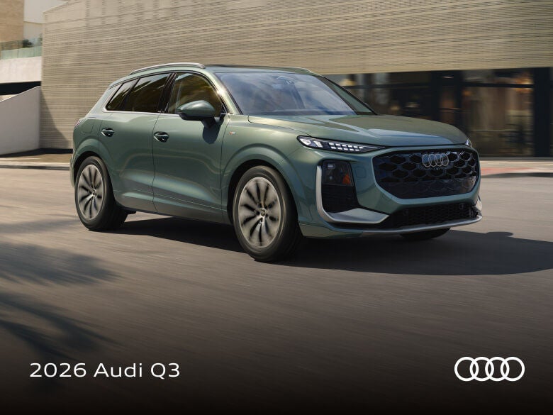 Front three-quarter view of a green 2026 Audi Q3 driving on a city street past a modern building.
