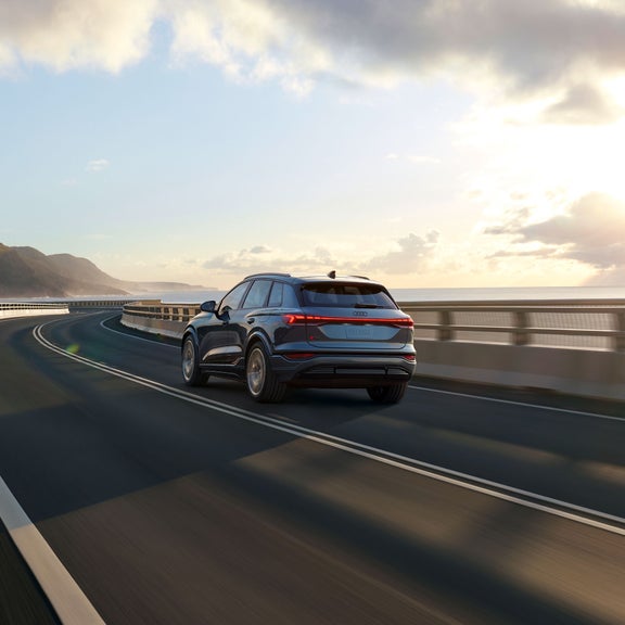 Three-quarter rear view of a blue Audi Q6 e-tron accelerating on an oceanside road.