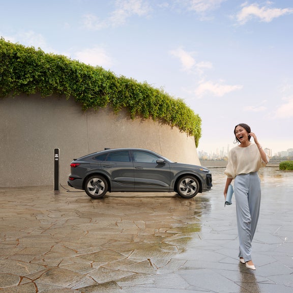 Side view of a grey Audi Q6 Sportback e-tron parked and charging with a person walking and laughing nearby.