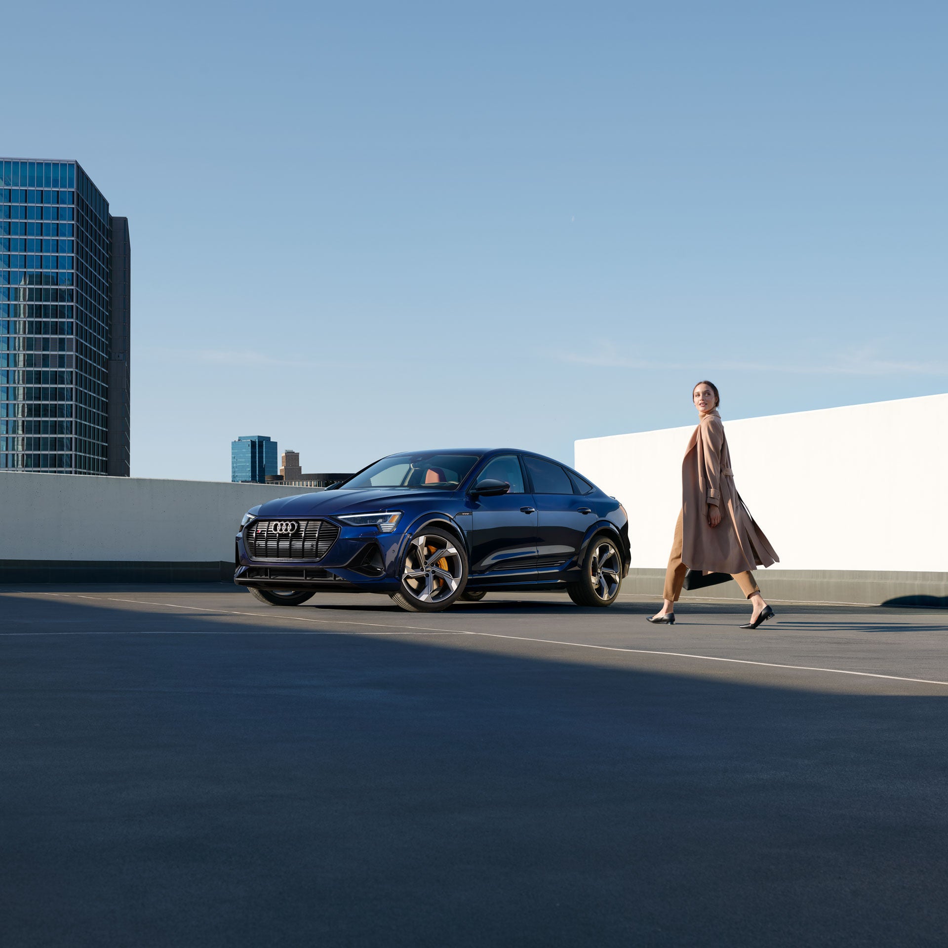 A woman walking towards an Audi e-tron in an empty parking lot with city buildings in the background.