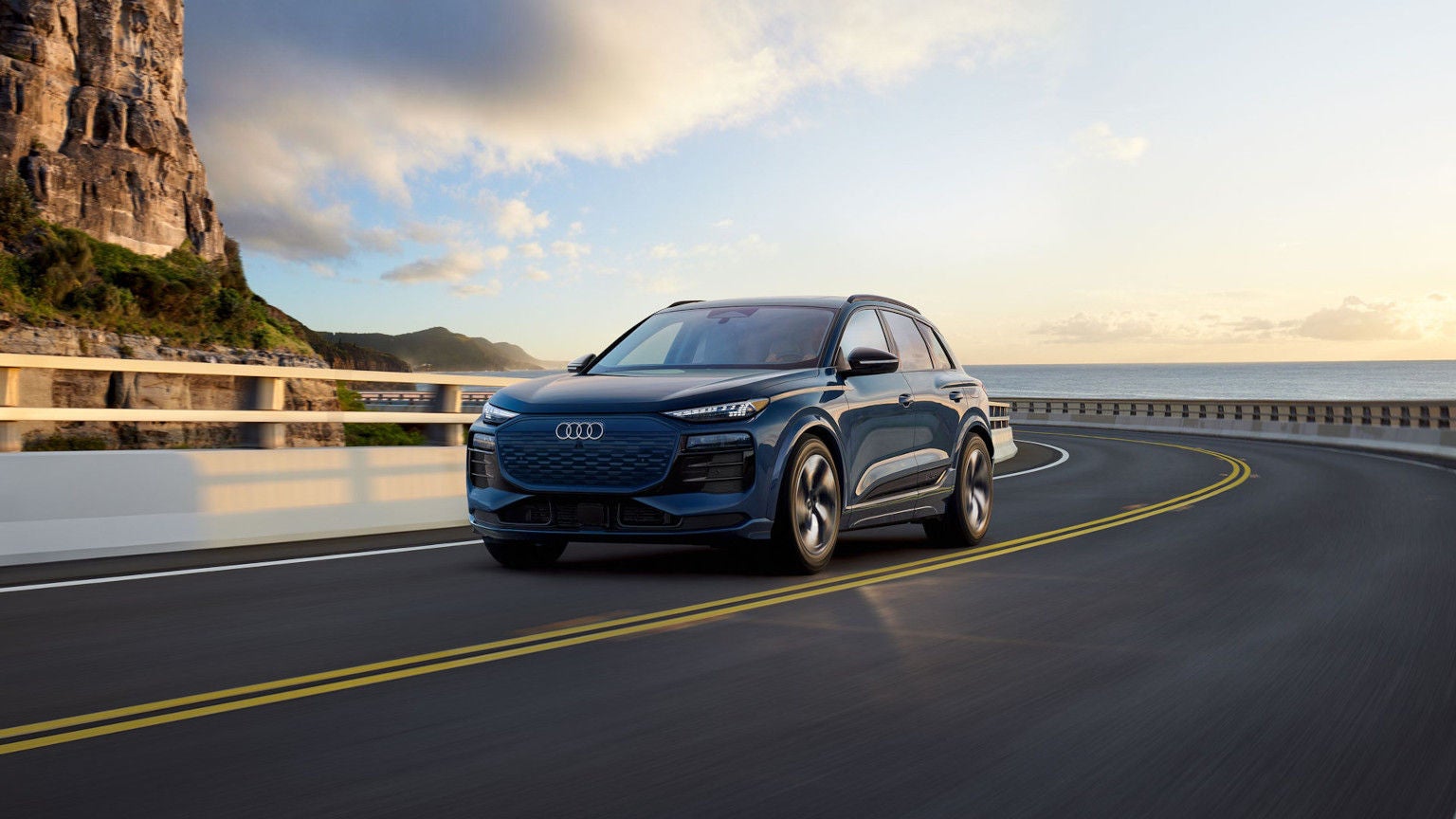 Image of a dark blue 2025 Audi Q6 e-tron driving on a winding oceanside road