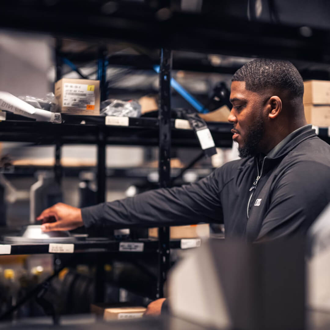 A man in a black Audi zip-up shirt grabs an Audi OEM auto part from a rack of auto parts.