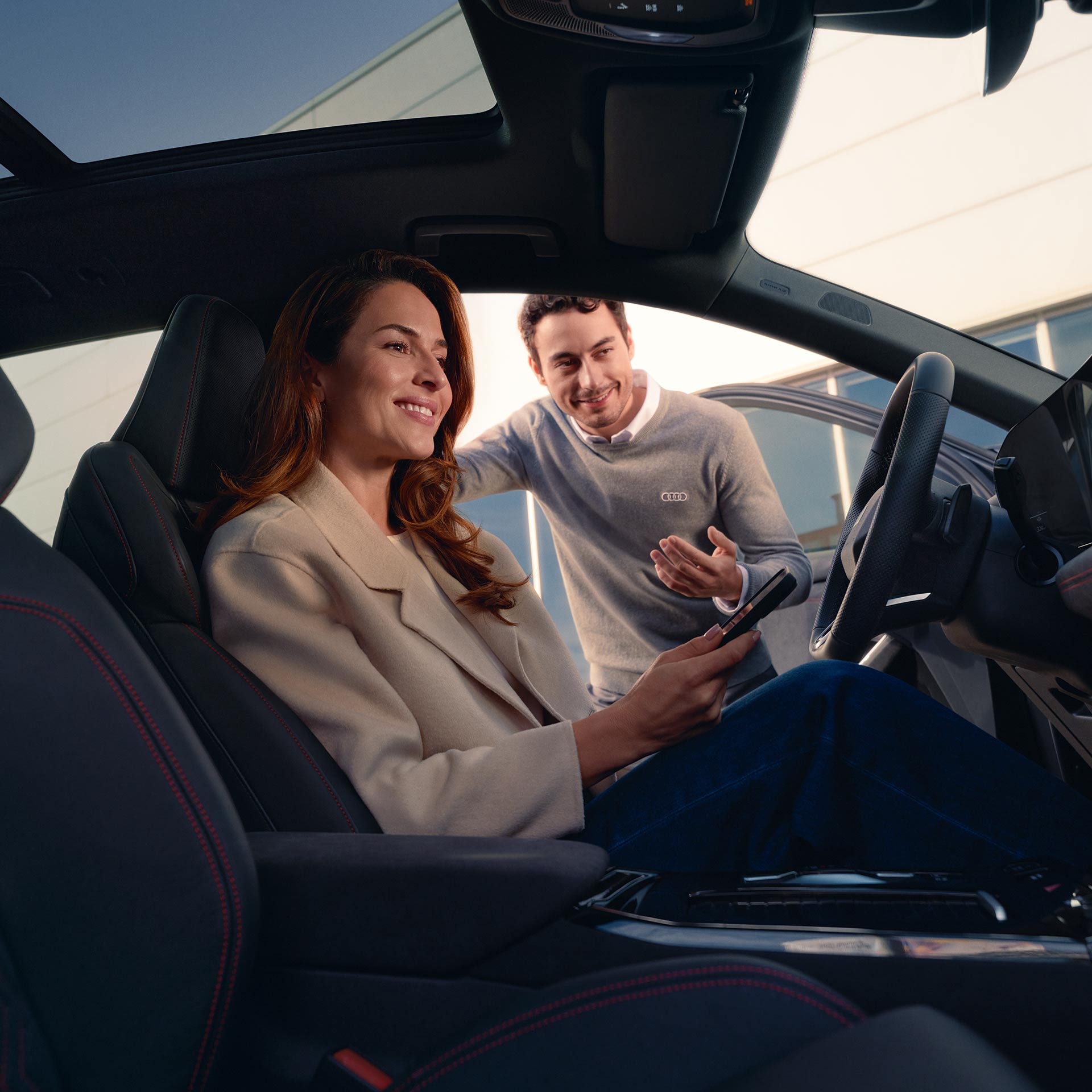 Interior view of an Audi vehicle showing an Audi customer in the driver seat and an Audi dealership representative leaning in the open driver's door.