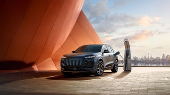 Three-quarter front view of the Audi SQ6 e-tron parked and charging by a modern building with a city landscape in the background.