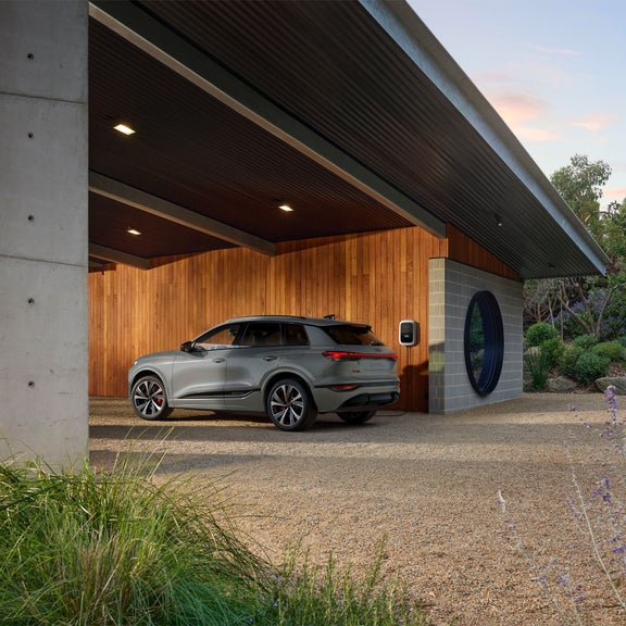 Plan latéral de trois quarts d’un Audi SQ6 e-tron gris en cours de recharge près d’un bâtiment moderne.