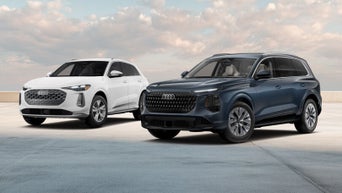 2026 Audi Q5 Premium in Arkona White and 2027 Audi Q7 Prestige in Alopias Blue Metallic, posed side by side under a soft cloudy sky.