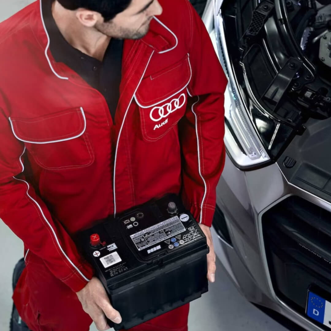 Overhead view of a man in a red Audi jumpsuit carrying an Audi OEM battery towards an Audi car with a popped hood.