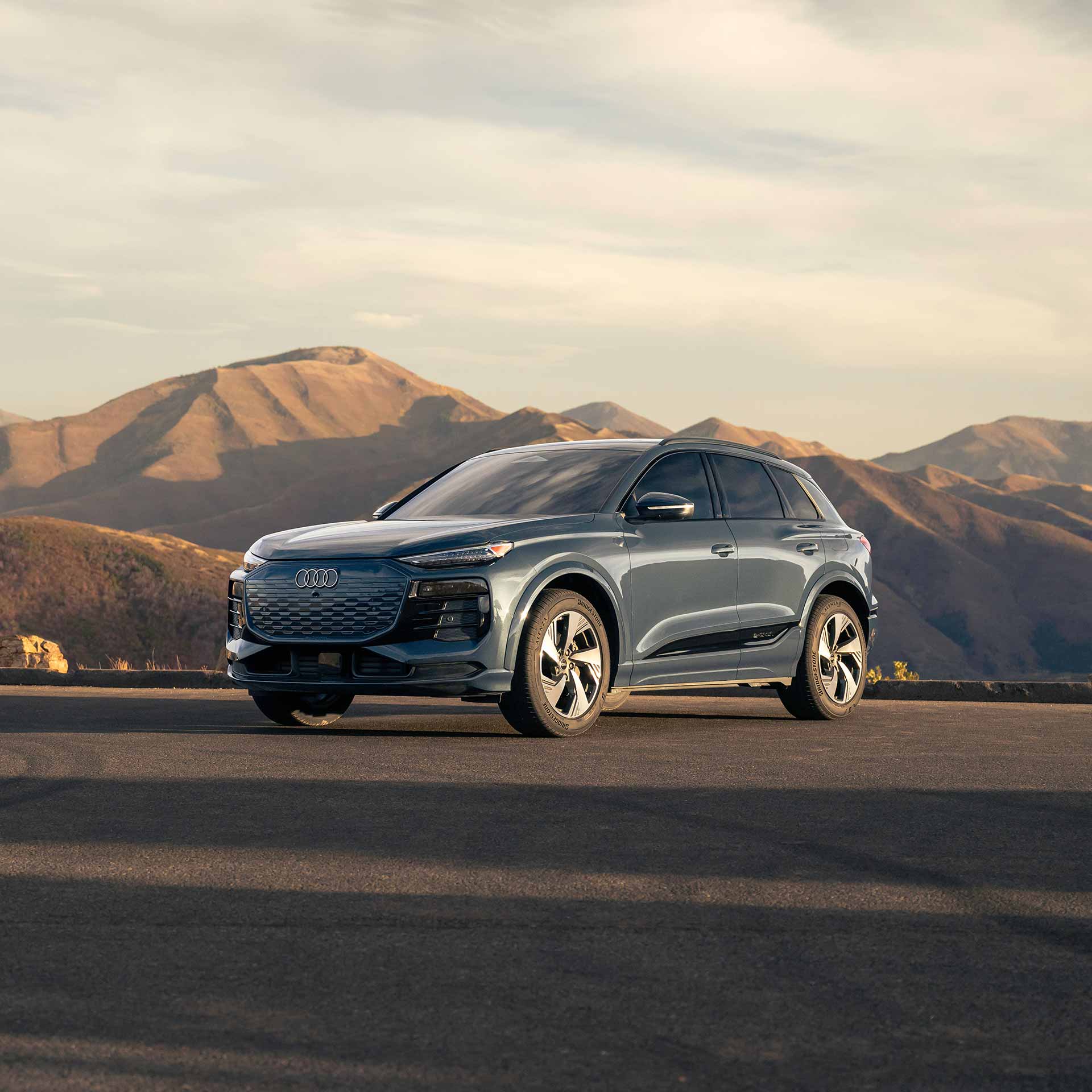 Three-quarter front view of a gray Audi A7 Sportback with illuminated headlights in front of a sunset.
