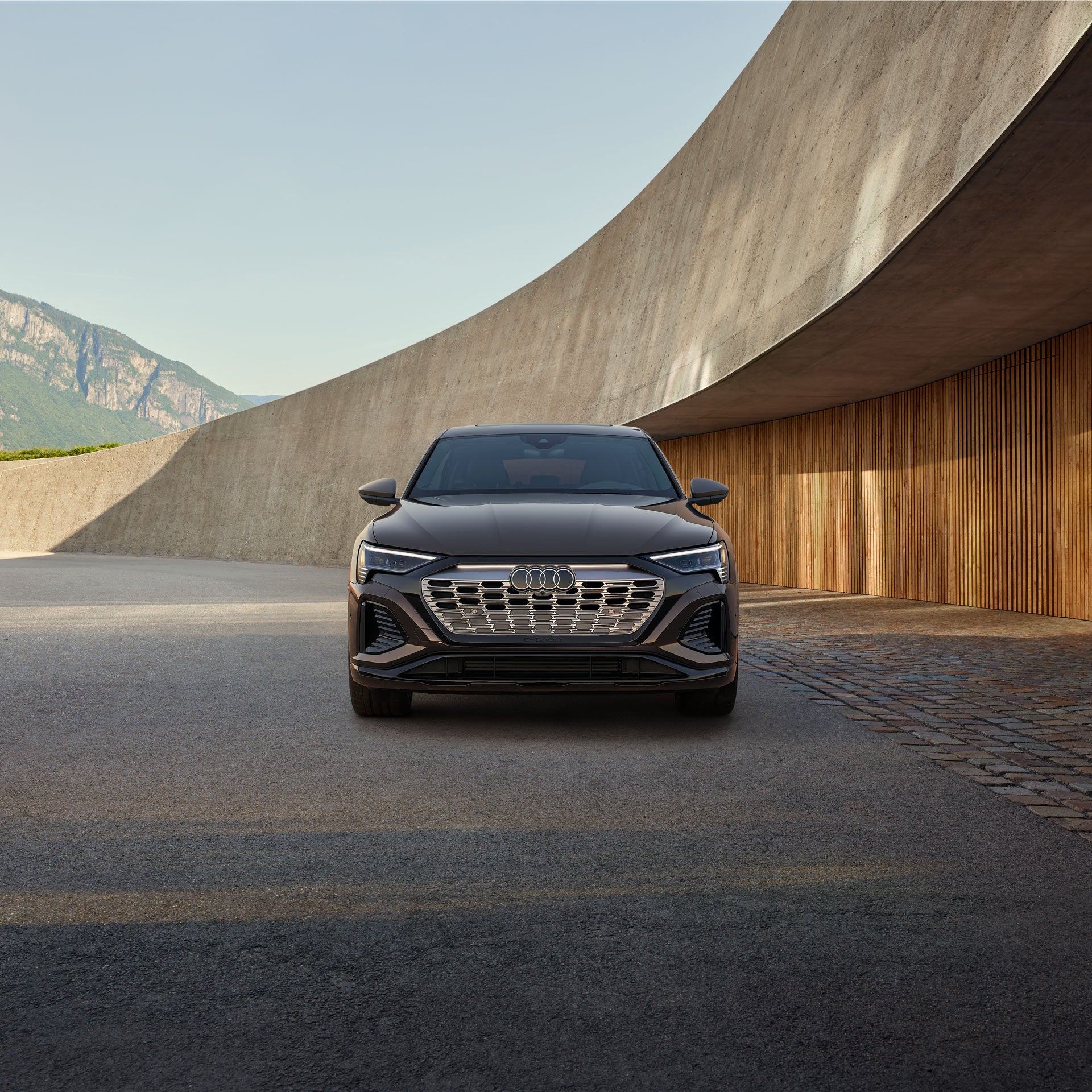 Front view of a brown Audi Q8 Sportback e-tron parked in front of a curved building with mountains in the background. 