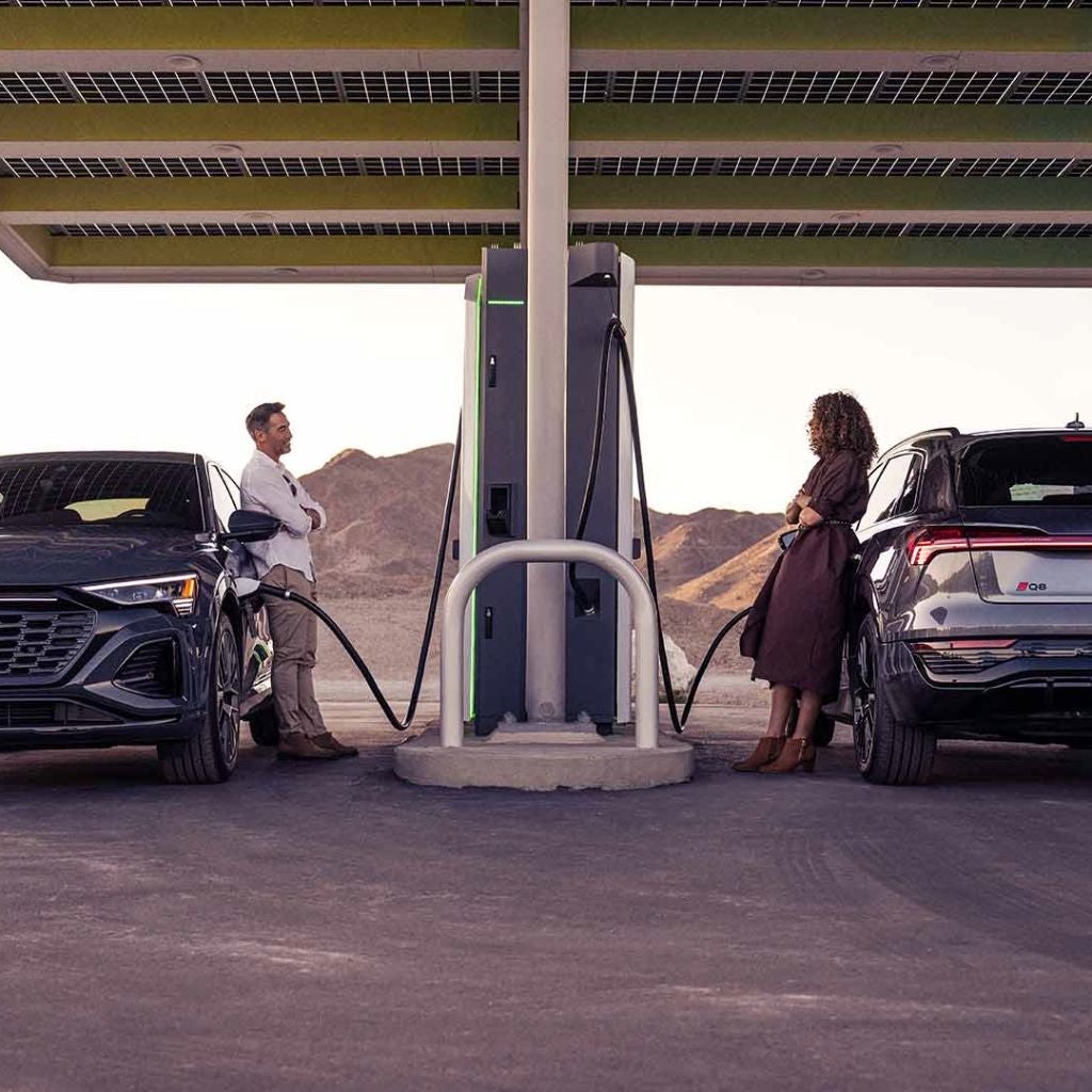 Two individuals leaning against their Audi vehicles, talking across the gas pump.