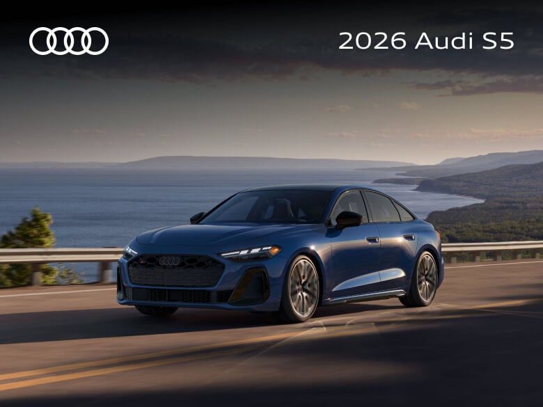 Three-quarter view of a blue 2026 Audi S5 driving on a coastal road overlooking the ocean.