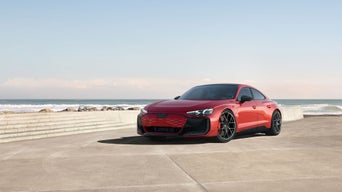 A silver Audi RS e-tron GT drives along a winding road at sunset, with glowing taillights and a mountain backdrop.
