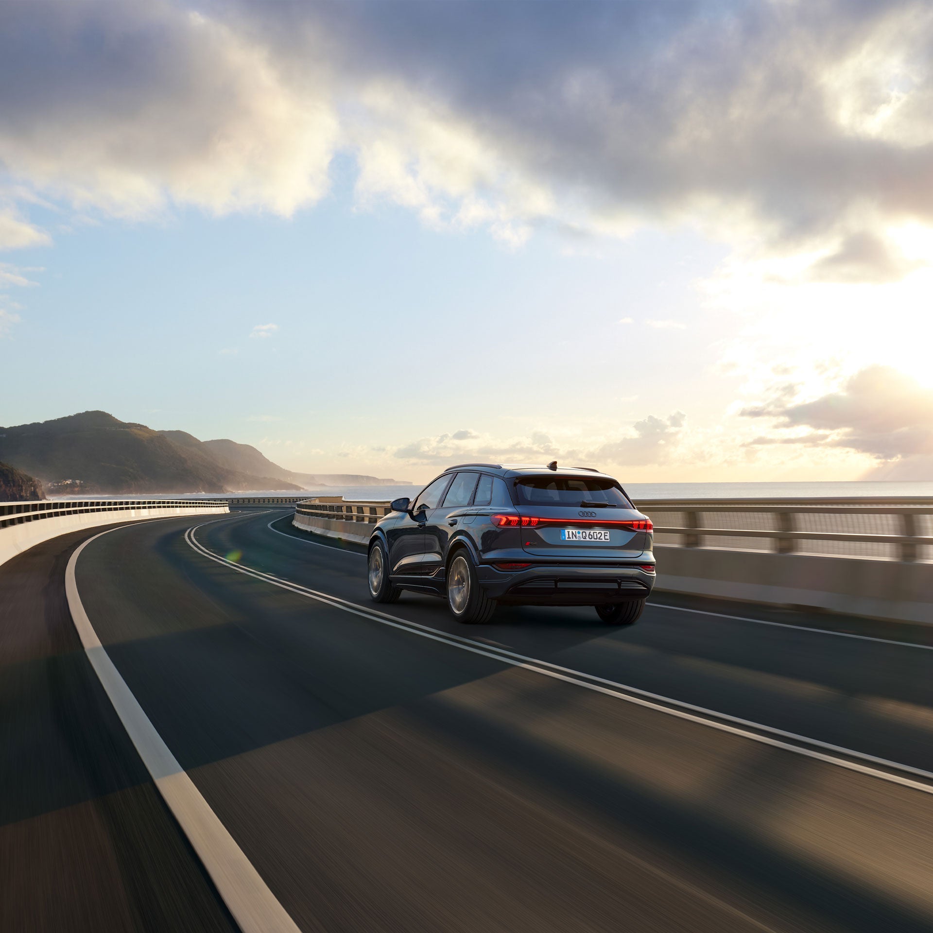 Three-quarter rear view of the Audi Q6 e-tron in motion along a coastal road.