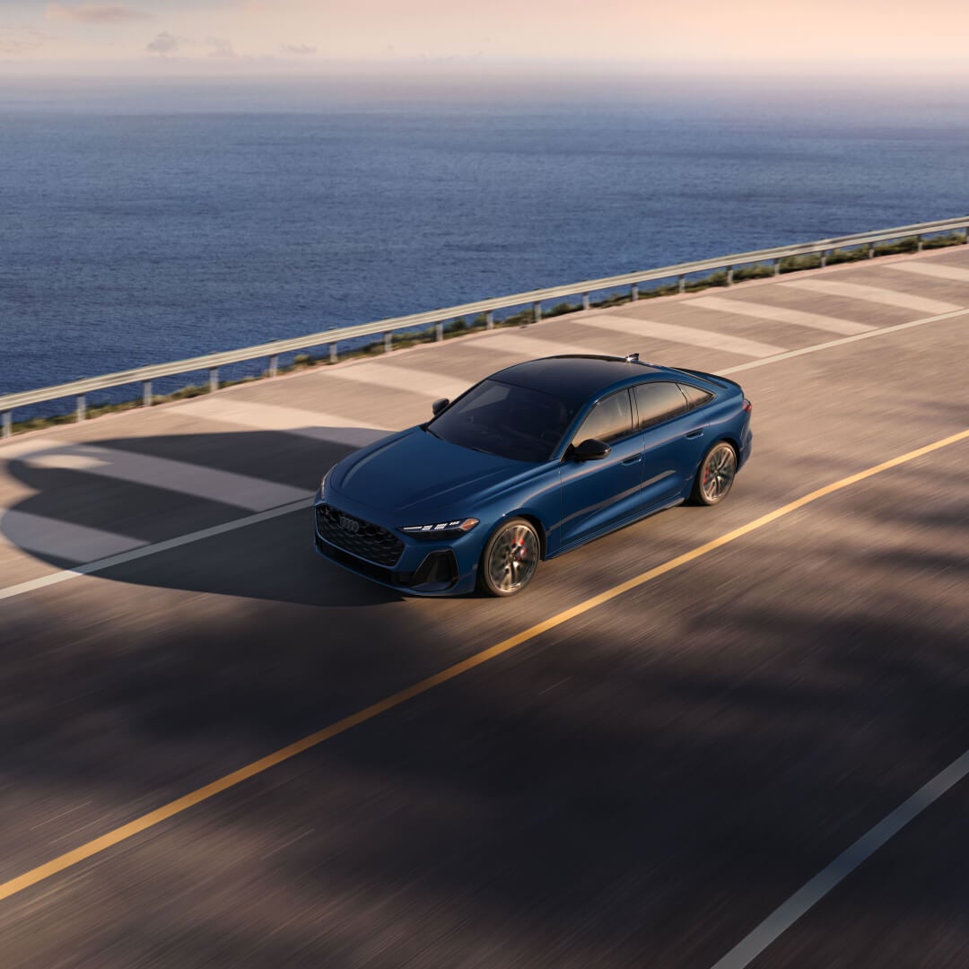 High-angle view of a blue 2026 Audi S5 cruising on a coastal highway by the ocean.
