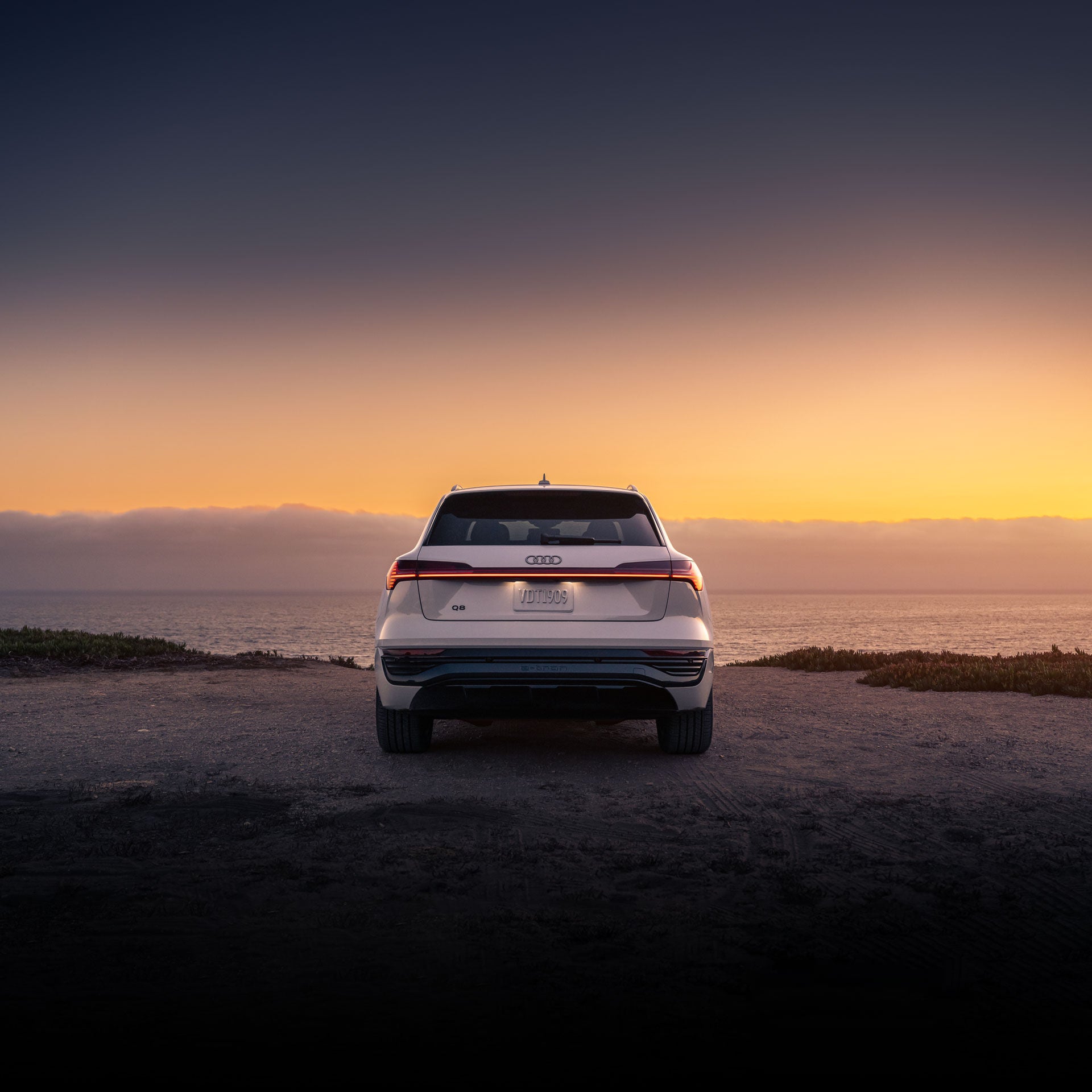 Rear shot of a white Audi Q8 e-tron parked in front of the ocean at sunset. 
